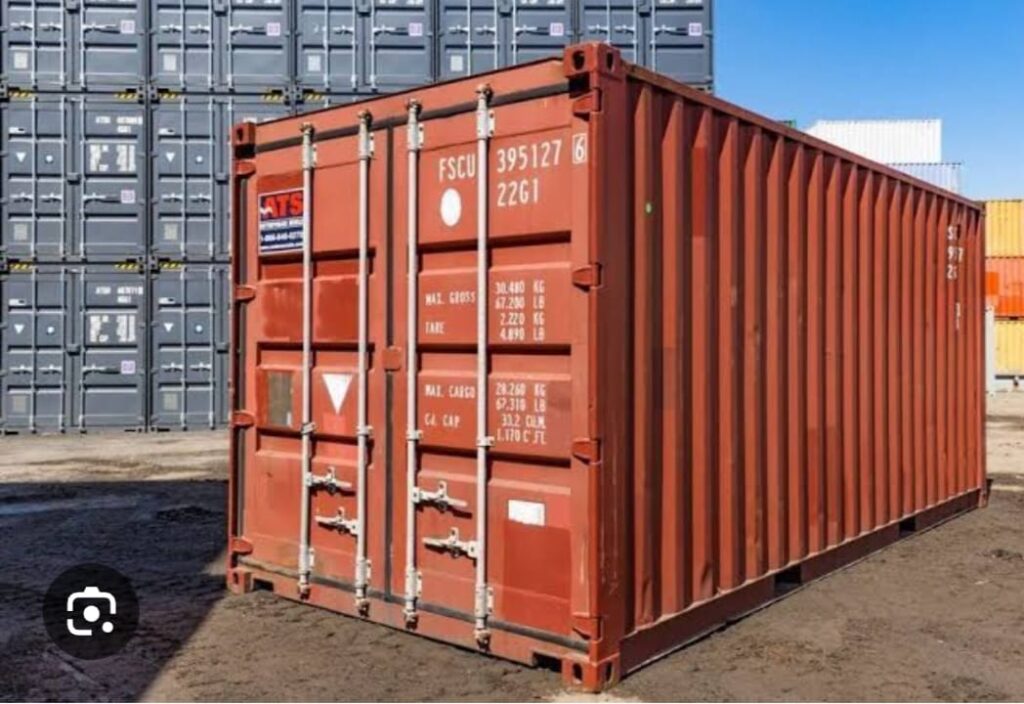 Red steel shipping container standing in a container yard with stacked cargo containers behind it.